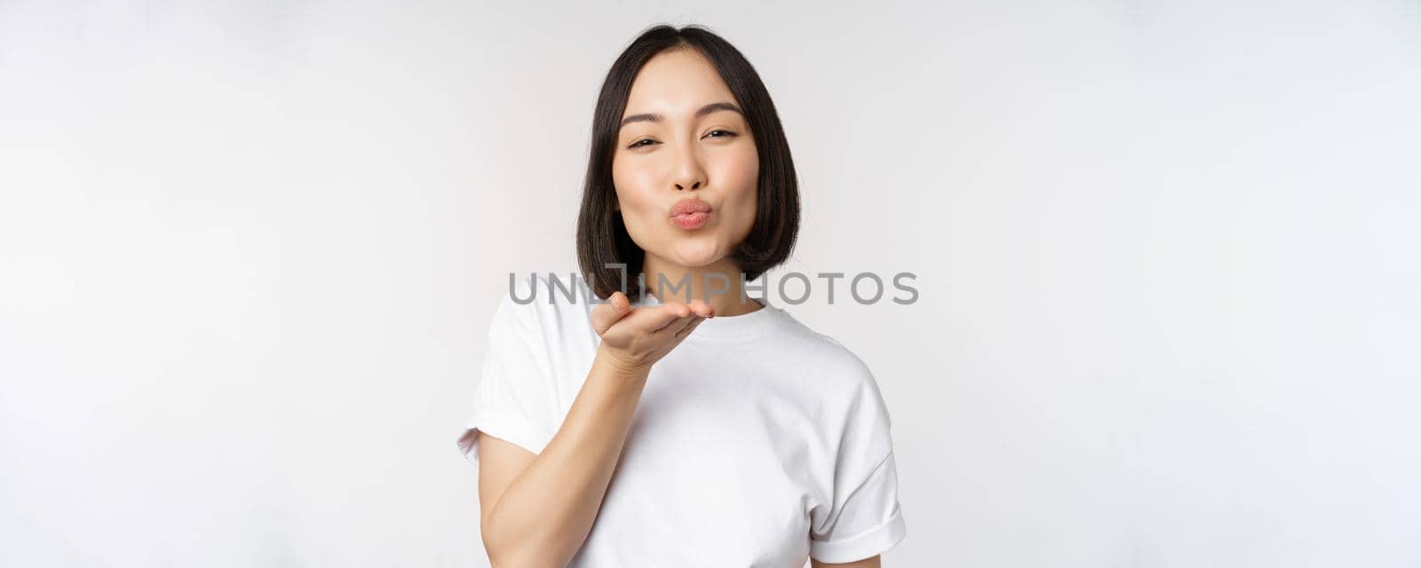 Lovely young asian woman, sending air kiss and looking coquettish at camera, standing in tshirt over white background by Benzoix