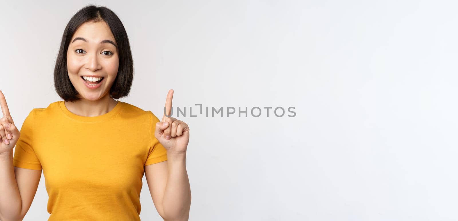 Happy beautiful asian girl showing advertisement, pointing fingers up, standing over white background by Benzoix