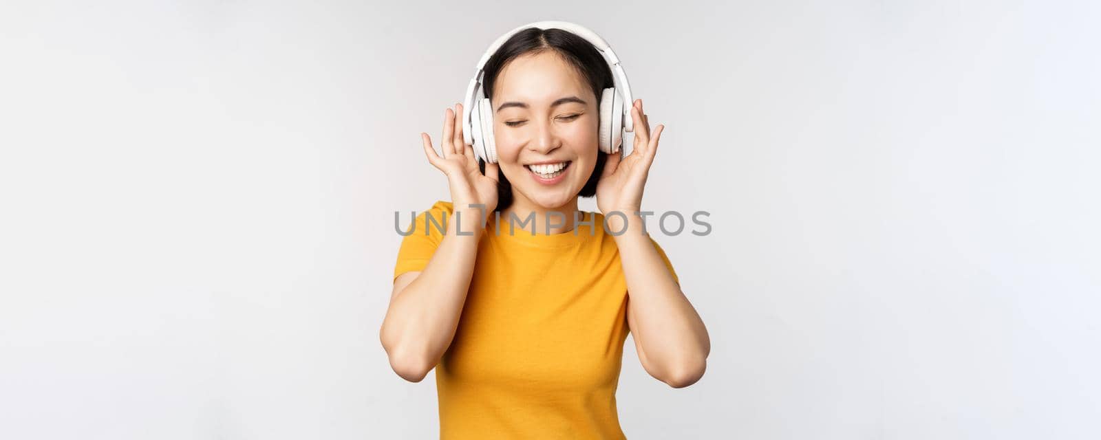 Happy asian girl dancing, listening music on headphones and smiling, standing in yellow tshirt against white background by Benzoix