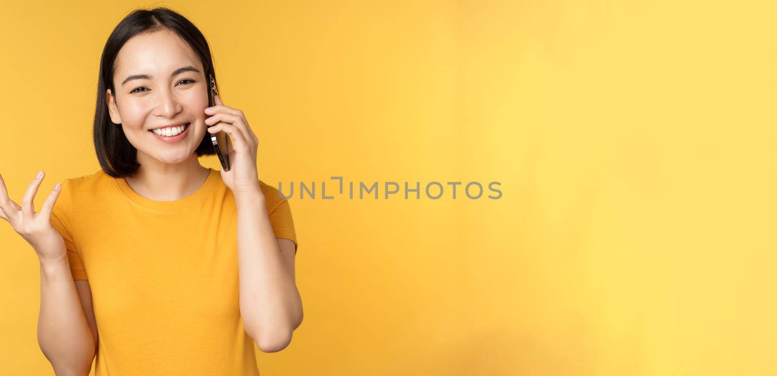 Young korean girl talking on mobile phone. Asian woman calling on smartphone, standing over yellow background.