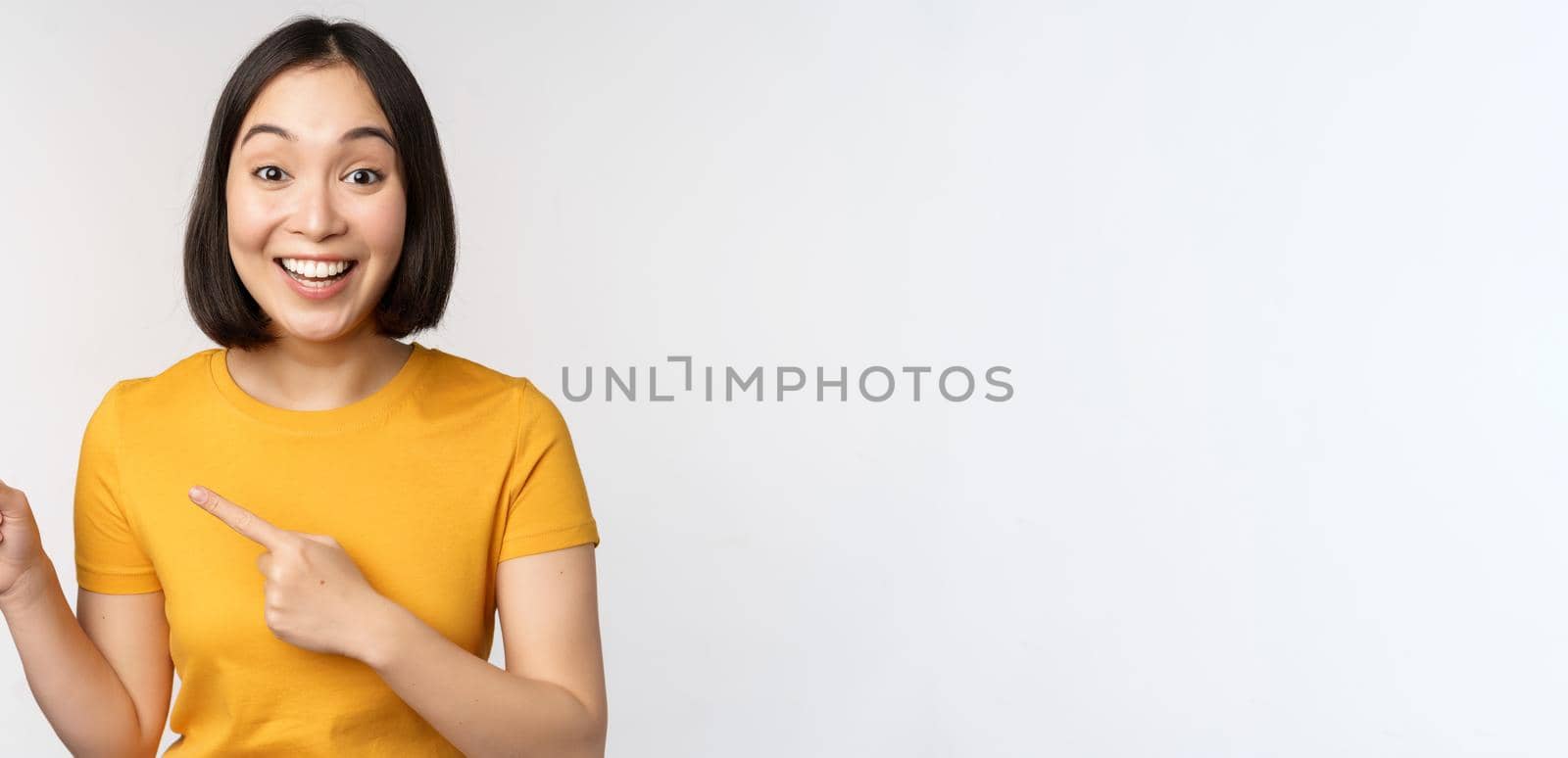 Portrait of smiling asian brunette girl in yellow tshirt, pointing fingers left, showing copy space, promo deal, demonstrating banner, standing over white background by Benzoix