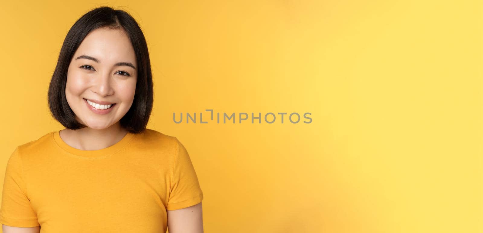 Close up portrait of beautiful asian woman smiling, looking cute and tender, standing against yellow background.