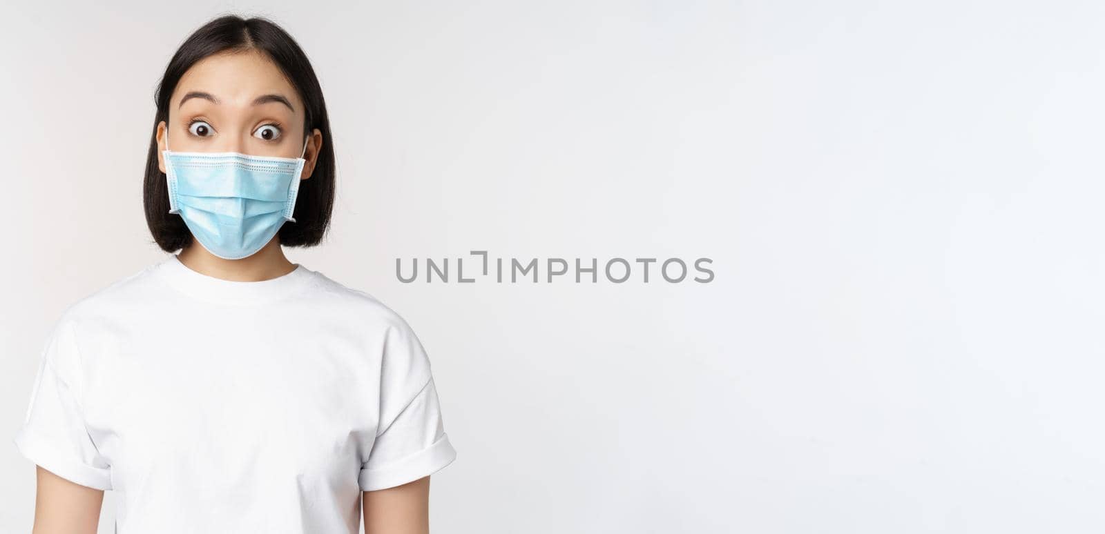 Health and covid pandemic concept. Image of korean woman in face medical mask looking surprised, reacting amazed, standing over white background by Benzoix