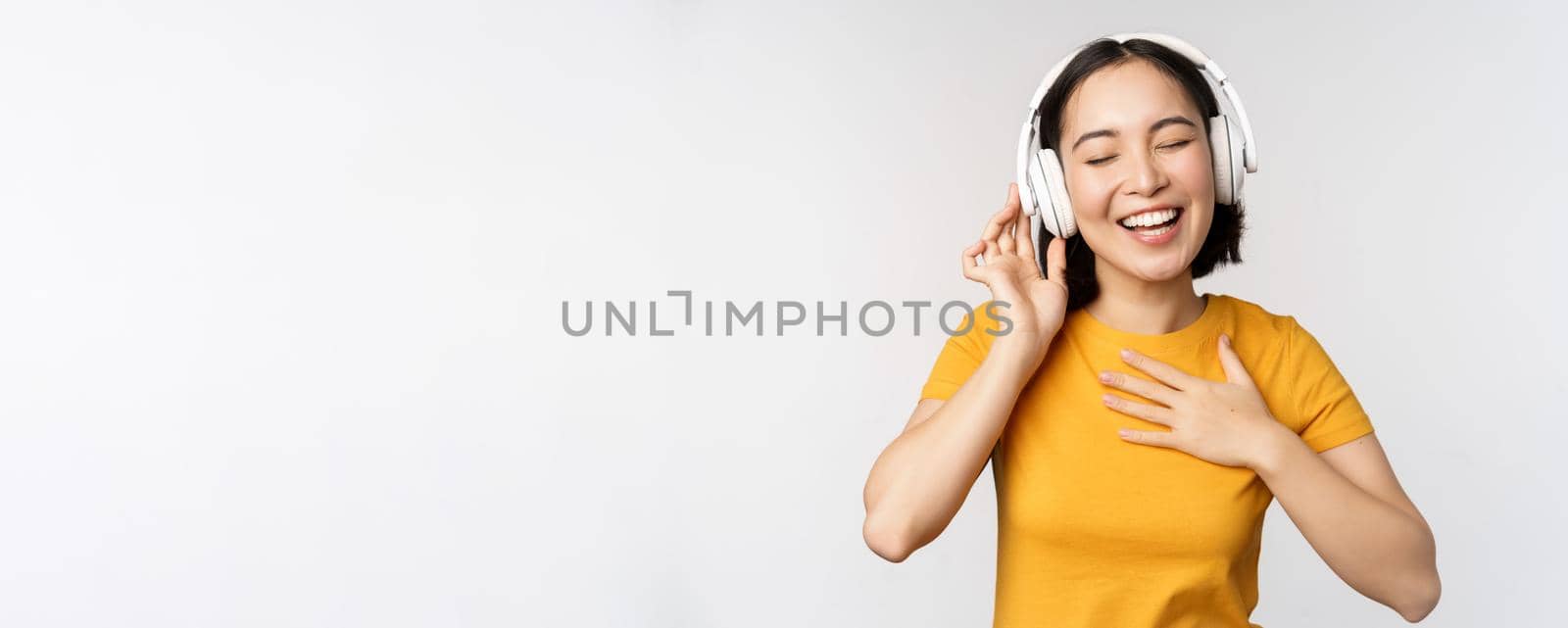 Happy asian girl dancing, listening music on headphones and smiling, standing in yellow tshirt against white background by Benzoix