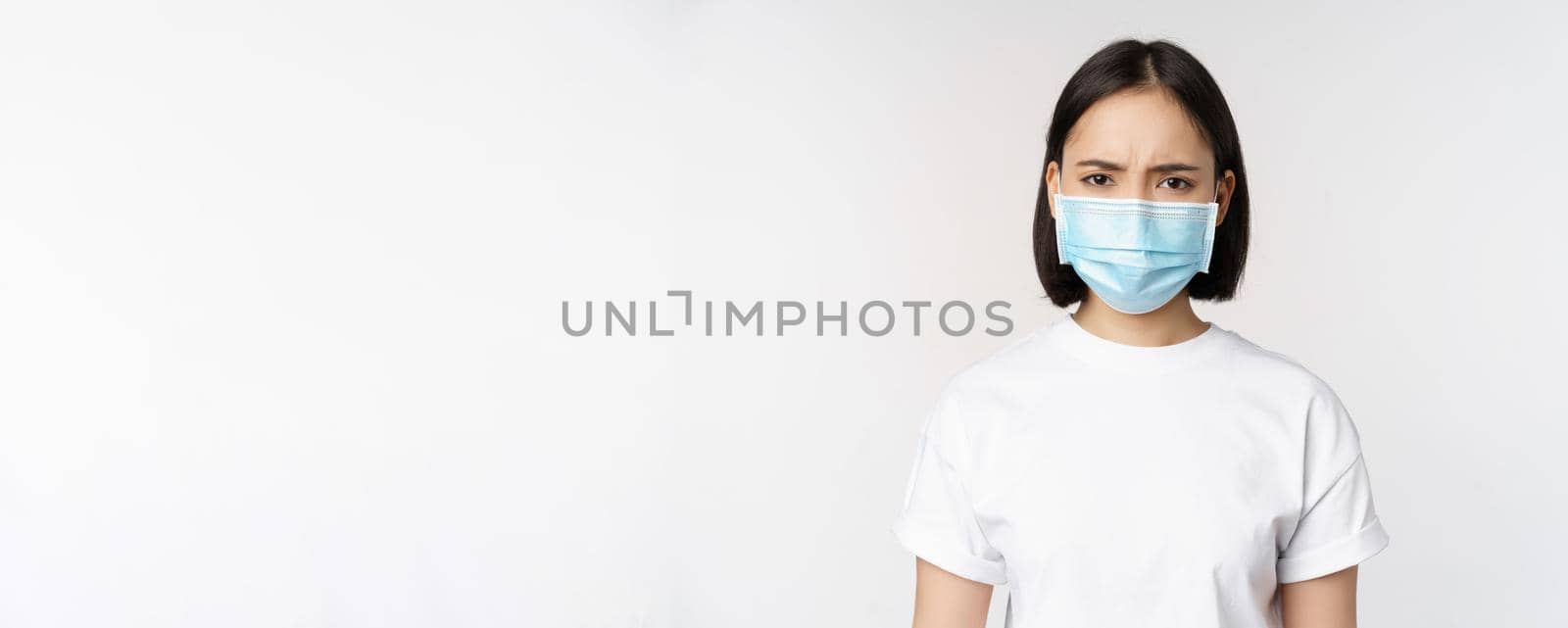 Health and covid pandemic concept. Angry young asian girl in face medical mask, frowning displeased, standing over white background.
