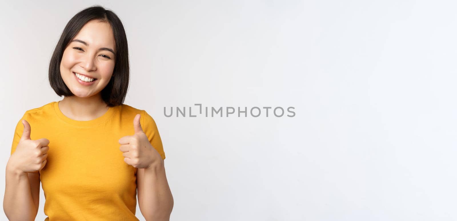 Beautiful smiling asian female model, showing thumbs up and looking pleased, recommending, express positive feedback, standing over white background by Benzoix