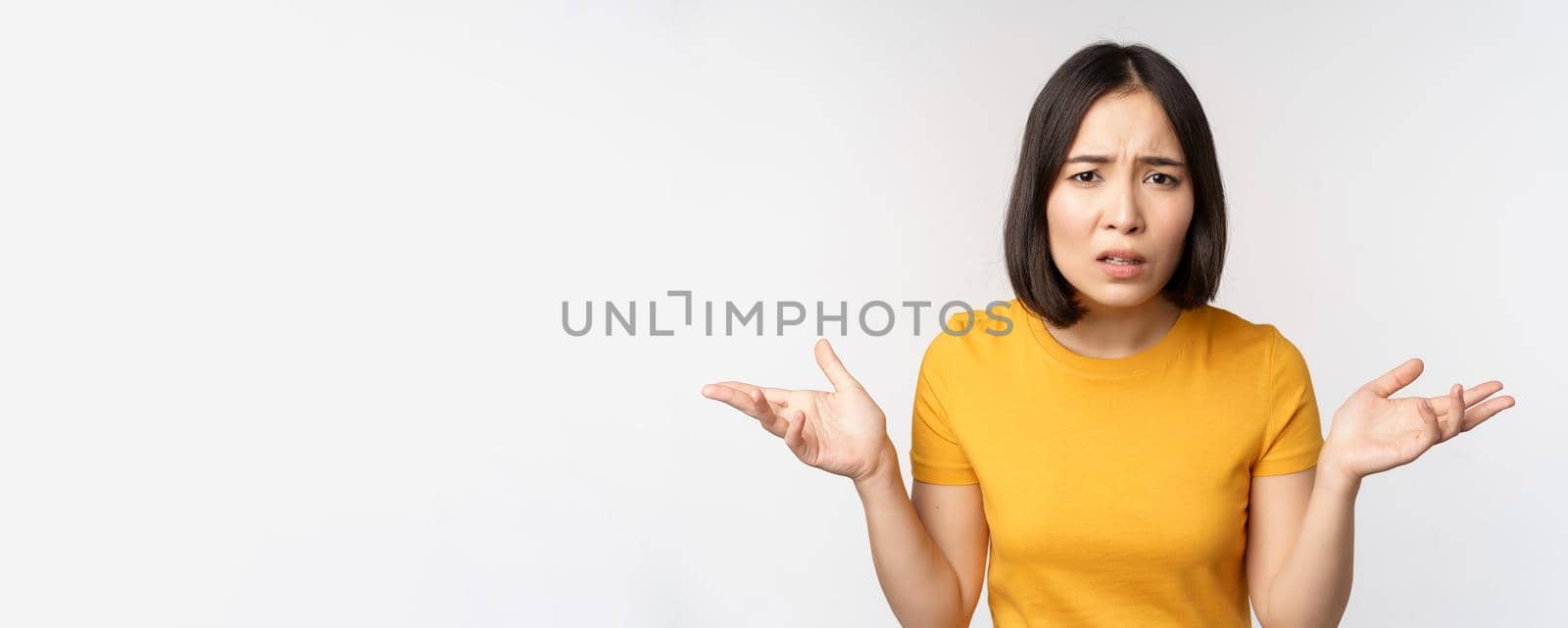 Frustrated, confused asian woman cant understand smth, looking clueless, shrugging shoulders, standing in yellow tshirt over white background.
