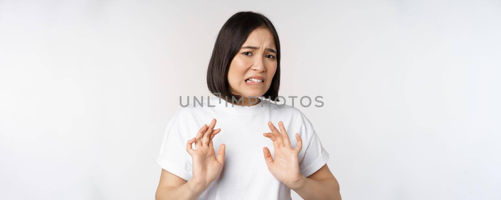 Disgusted asian woman rejecting smth, grimacing from dislike and aversion, stare with cringe, refusing offer, standing over white background.