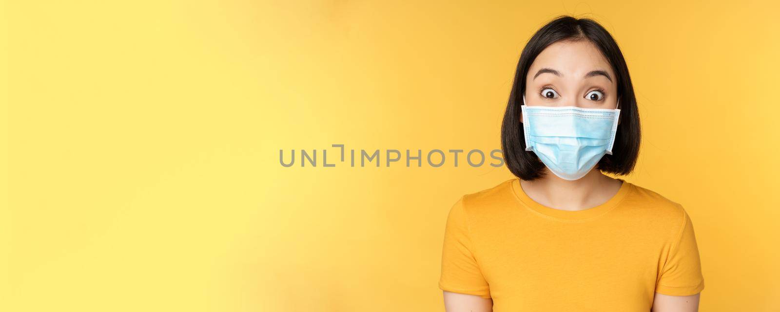 Covid-19 and medical concept. Close up portrait of asian woman in face mask, looking surprised and amazed at news, standing over yellow background by Benzoix