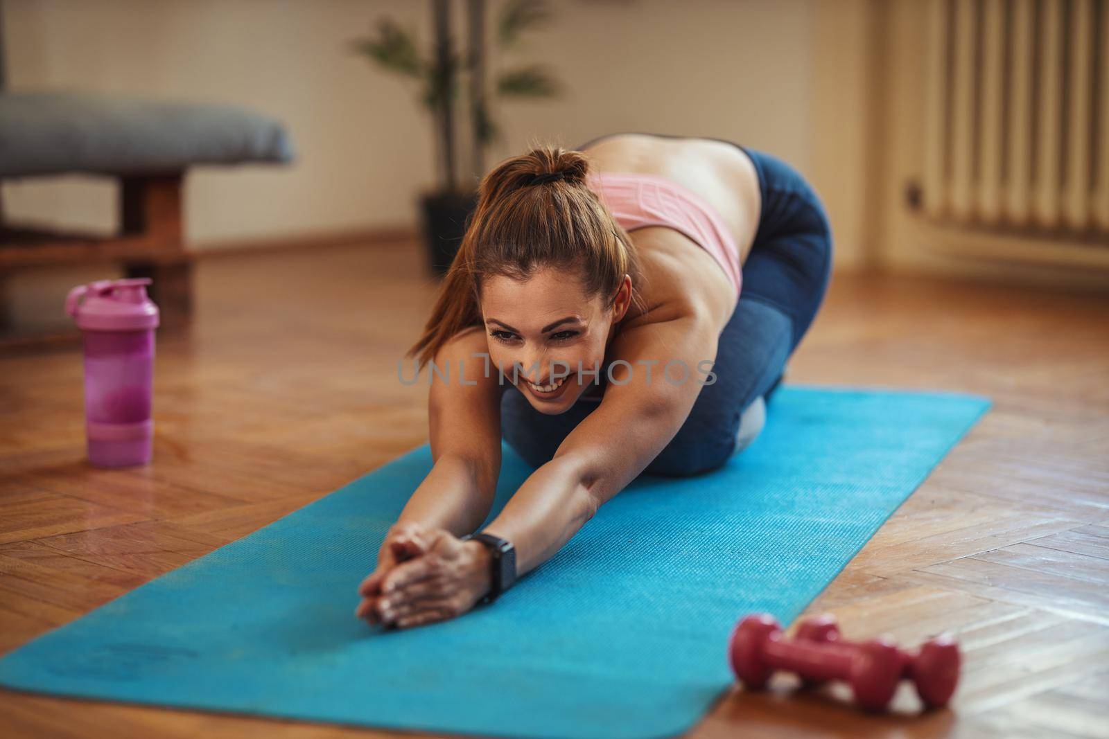 Young smiling woman is doing stretching exercises in the living room on floor mat at home in morning sunshine.