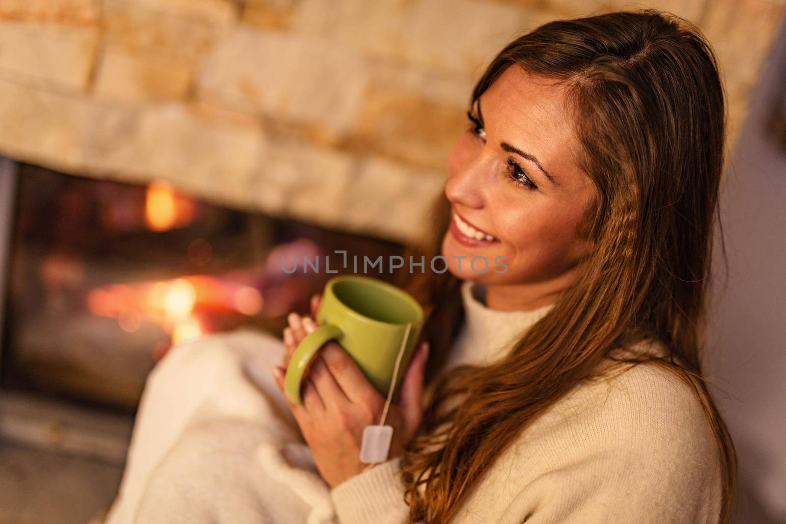 Beautiful young smiling woman enjoying a cup of tea by the fireplace.