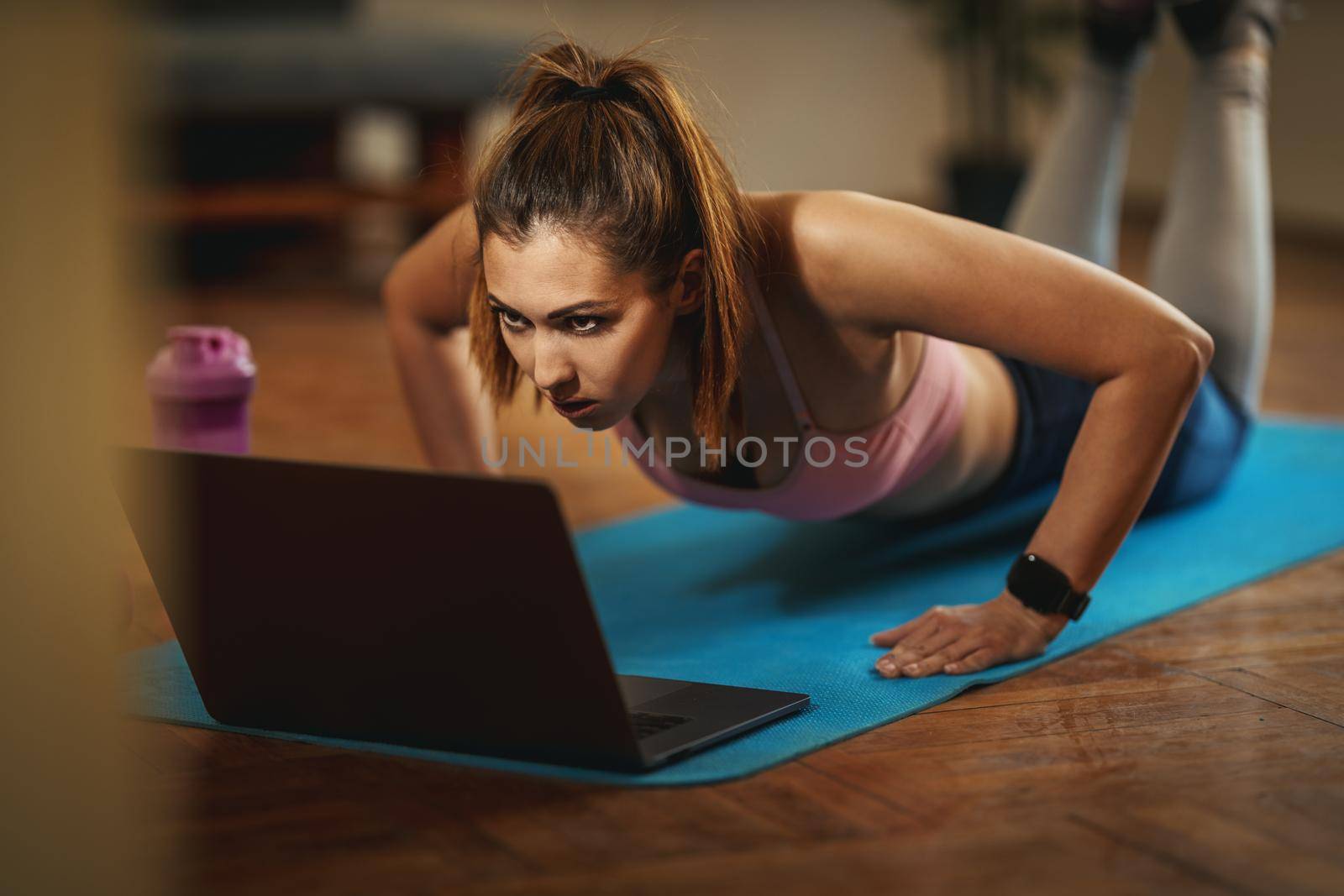 Young smiling woman is doing fitness exercises in the living room on floor mat at home and looking at the laptop.
