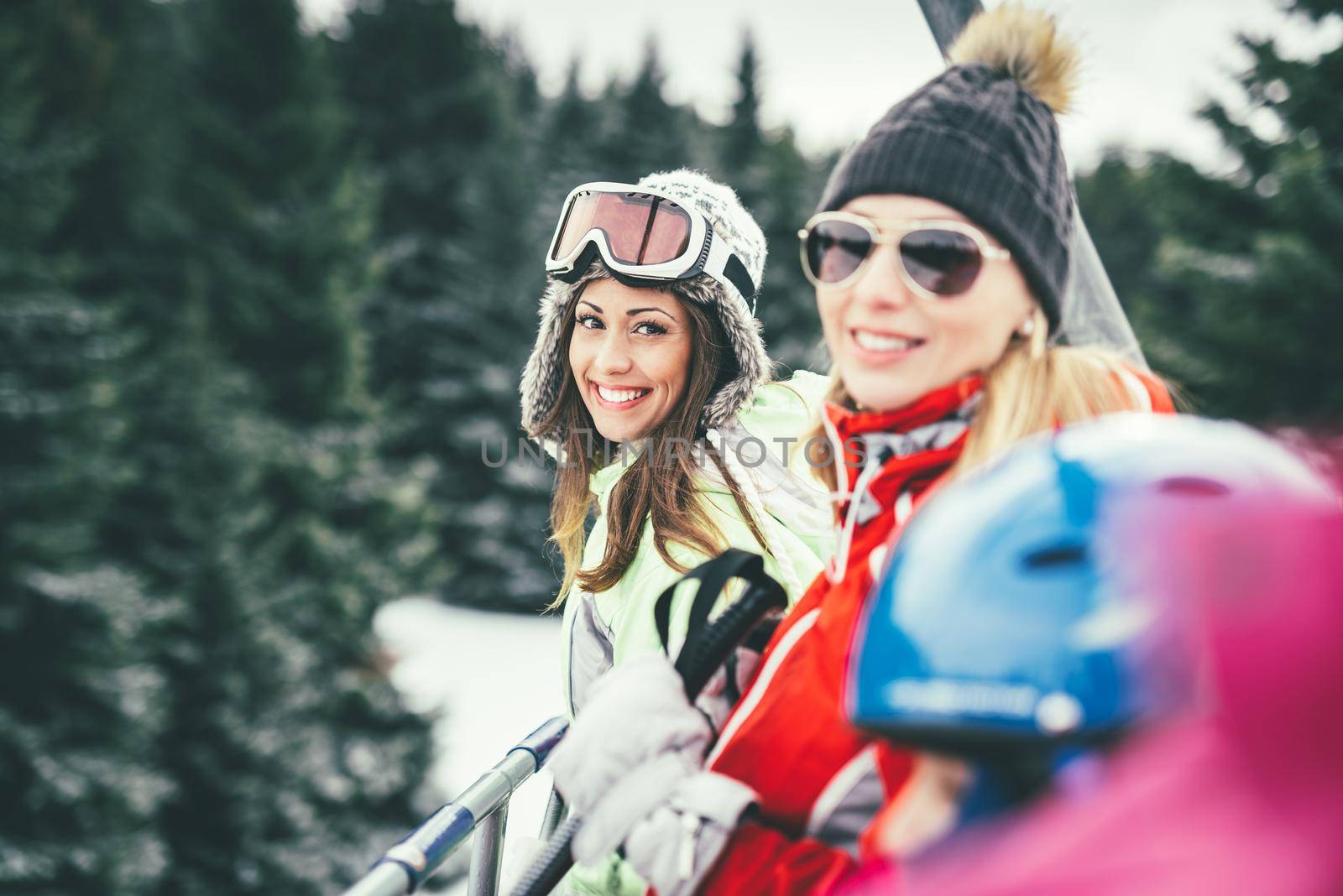 Beautiful young woman friends enjoying in winter vacations. They driving on ski lift and looking at camera with smile.