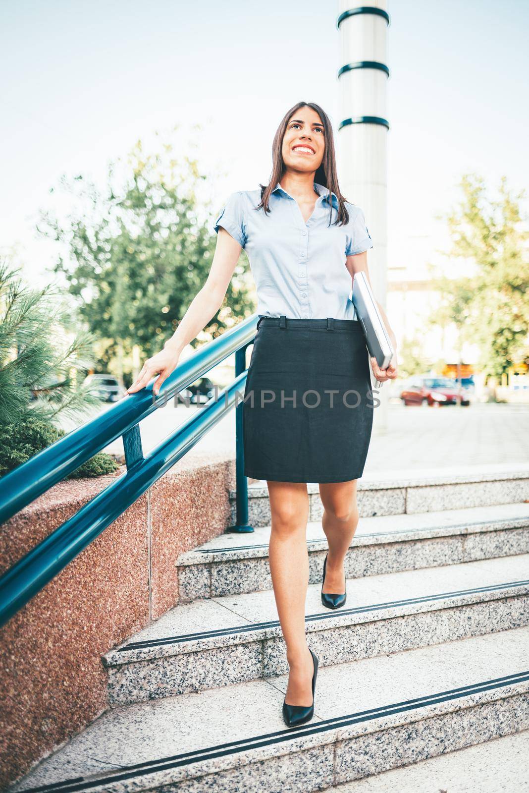Young businesswoman is walking down stairs to the building. She is holding laptop and goes to work.