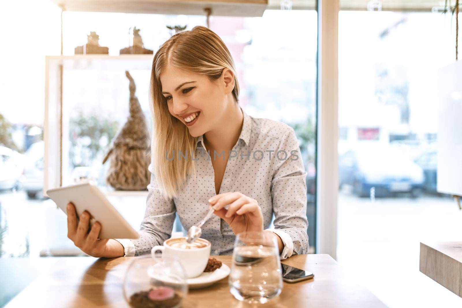 Beautiful young smiling woman drinking coffee in a cafe. She is surfing the internet on a digital tablet.