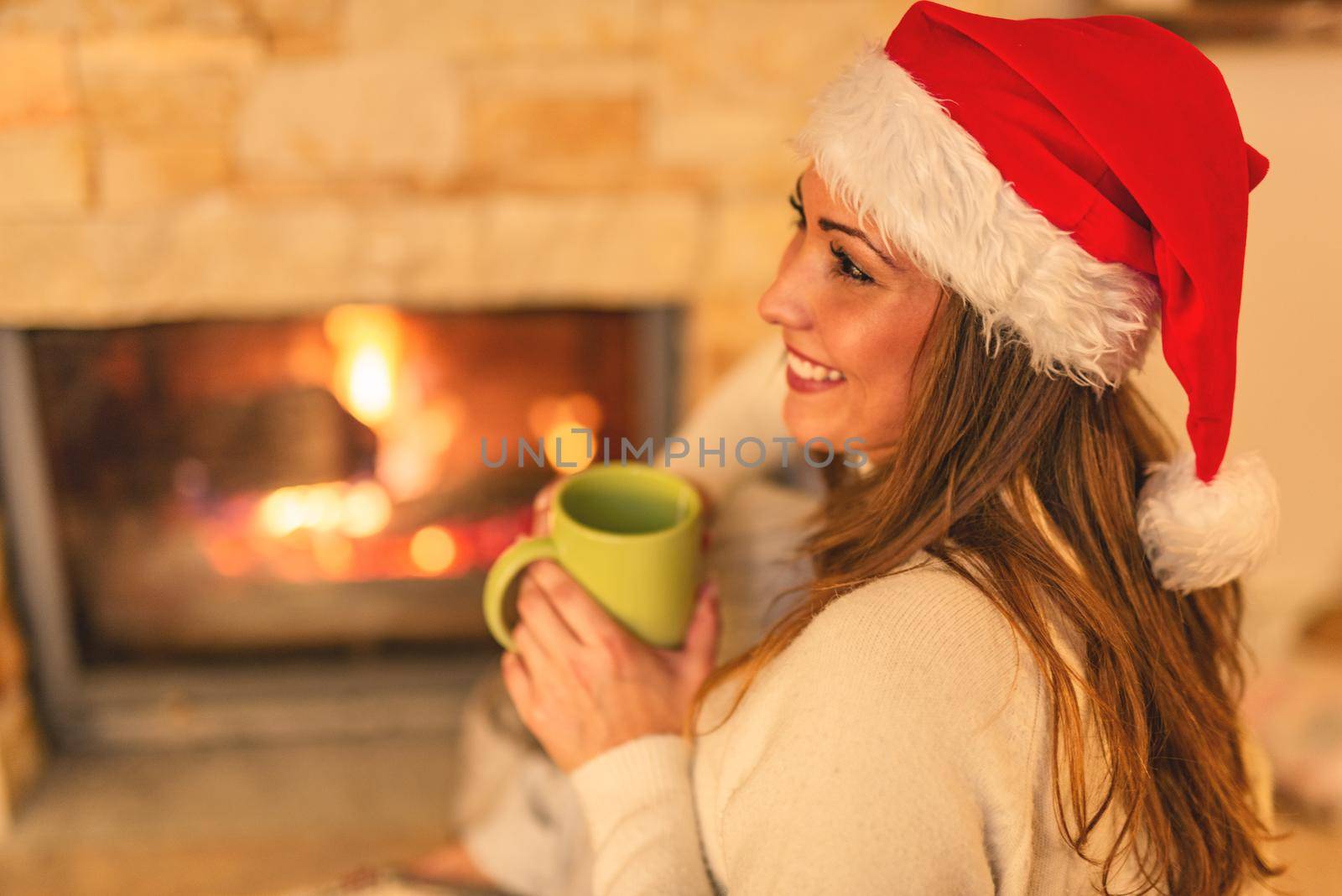 Beautiful young smiling woman enjoying a cup of tea by the fireplace in Christmas evening.