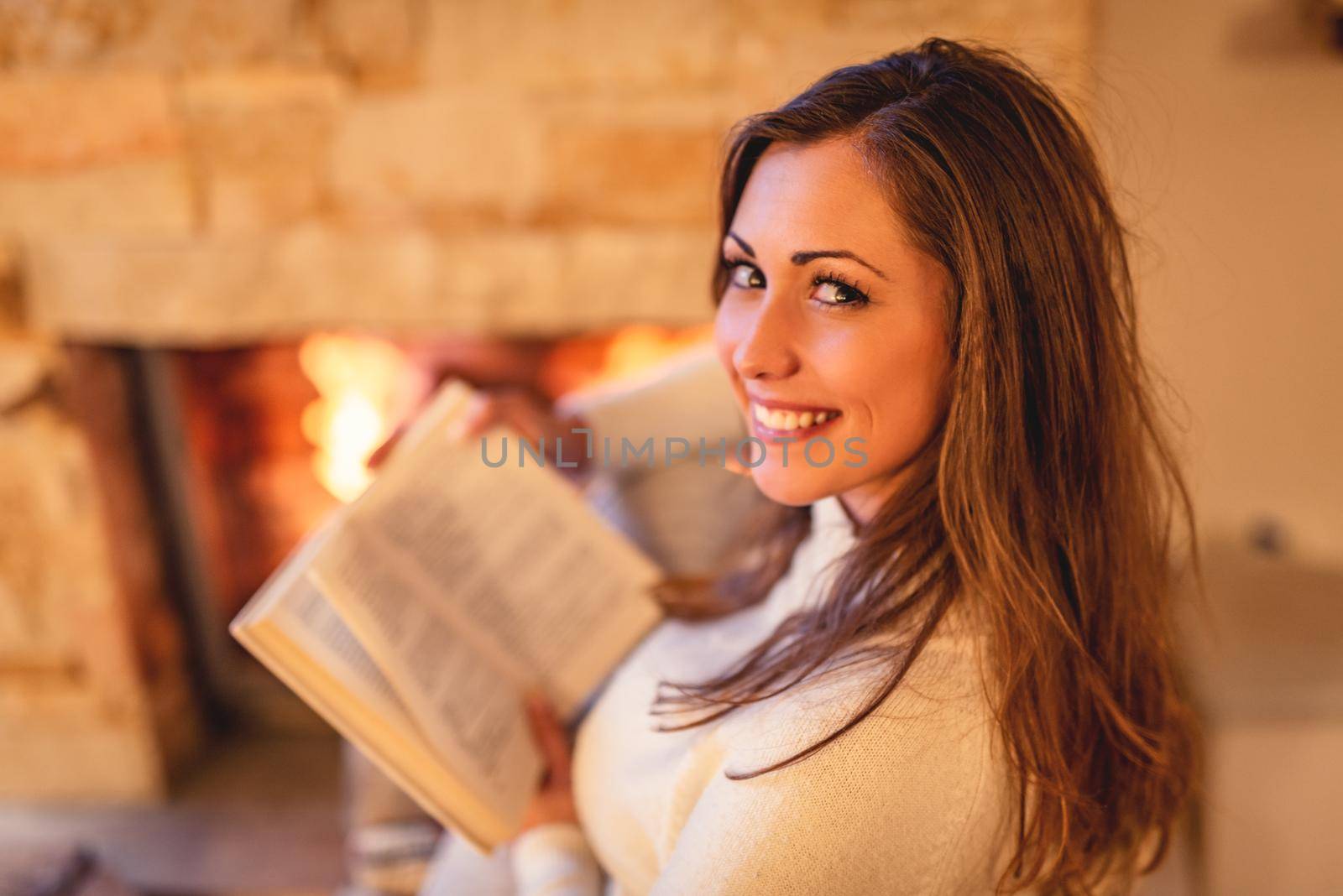 Beautiful young smiling woman reading book and enjoying by the fireplace.