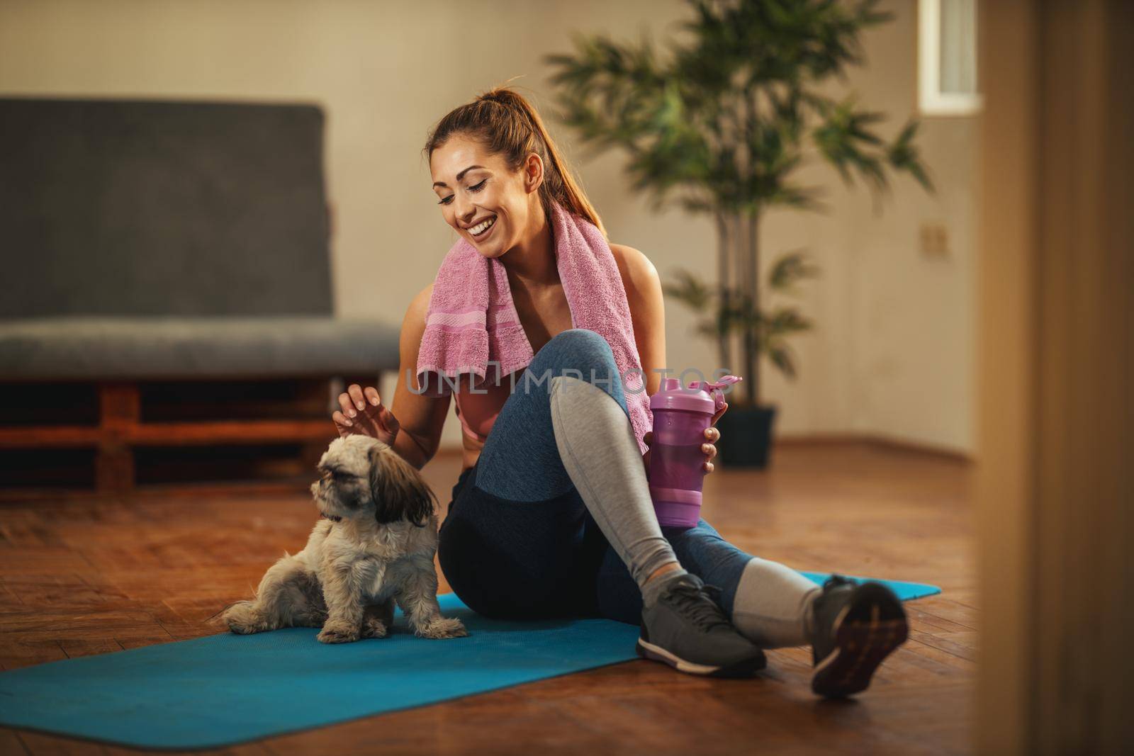 Young woman is doing stretching exercises in the living room at home supporting by her pet dog. She is resting on floor mat in morning sunshine.