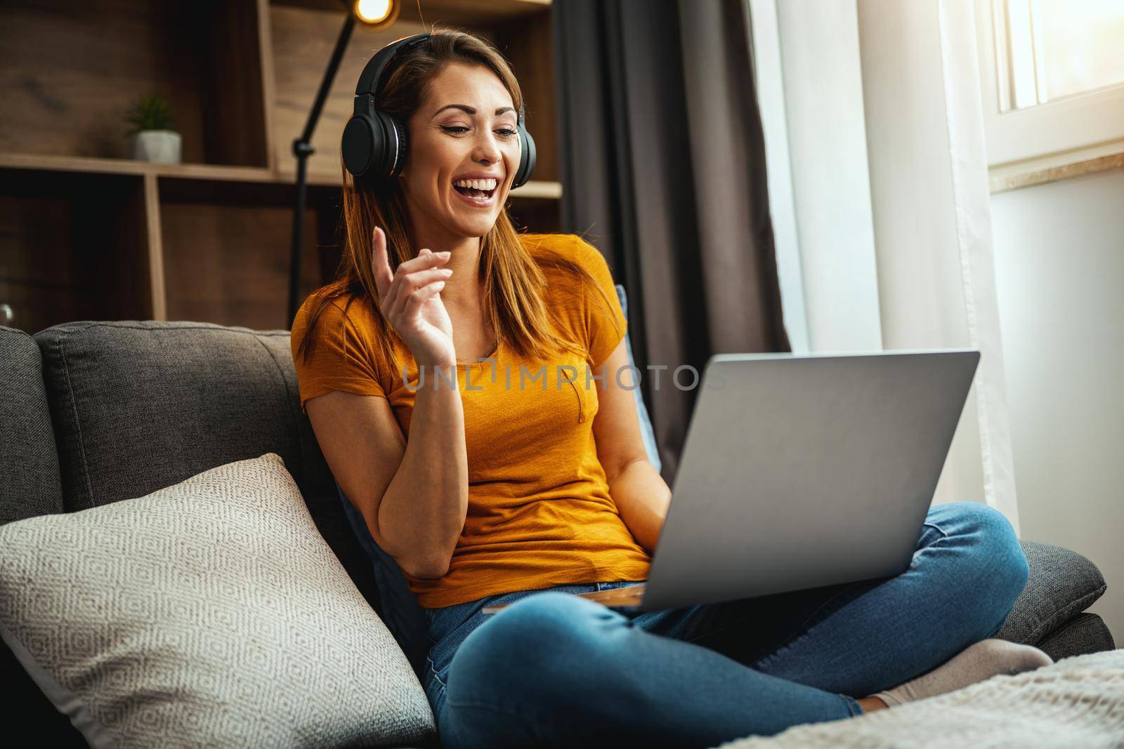 Attractive young woman sitting cross legged on the sofa and using her laptop and headphones to make a video call with someone at home.