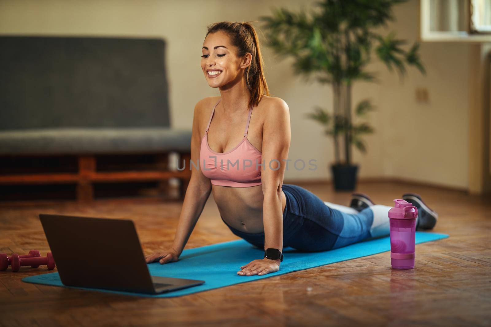 Young smiling woman is doing stretching exercises in the living room on floor mat at home, looking at the laptop, in morning sunshine.