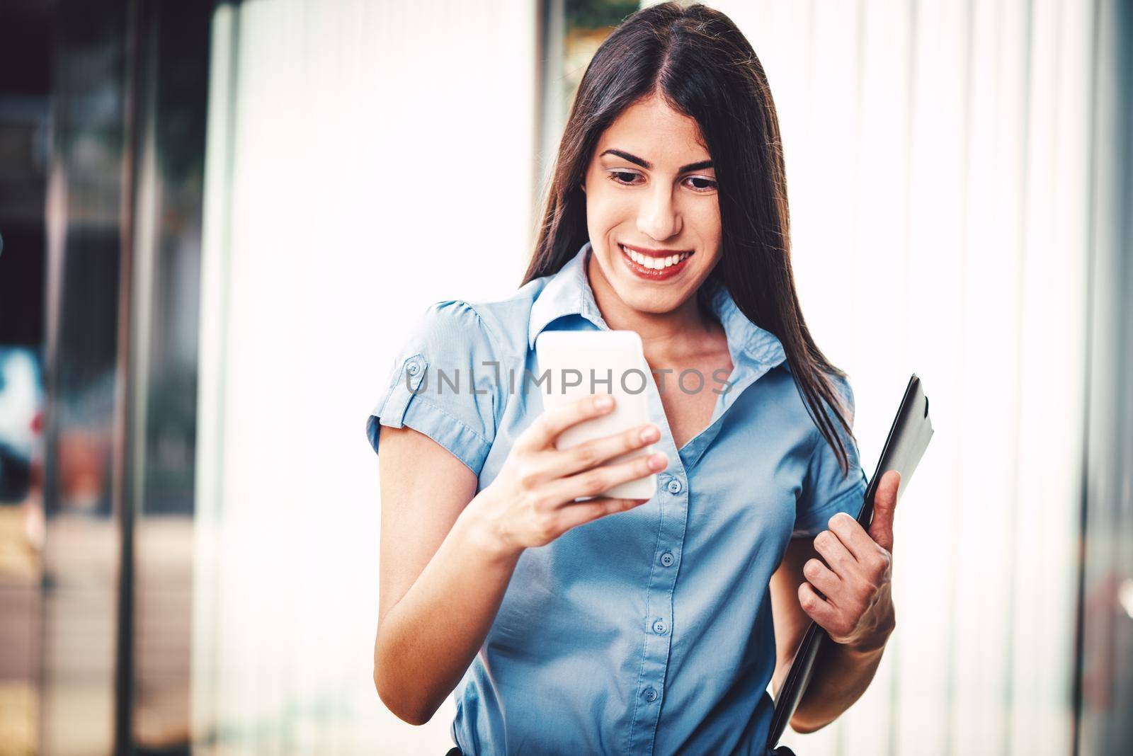 Emotional portrait of a happy and beautiful young businesswoman texting on a smartphone and carries a file document on the background of a business center.
