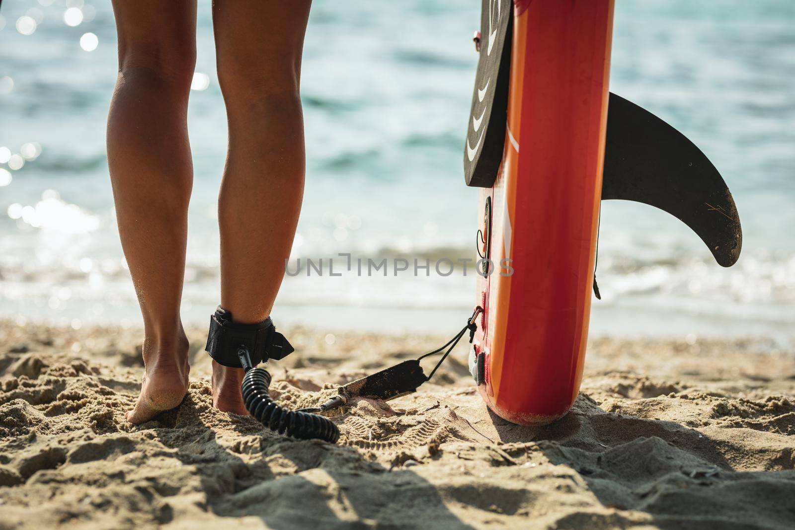 Close-up of young woman's legs on a sea beach and a surfboard, ready for the waves.
