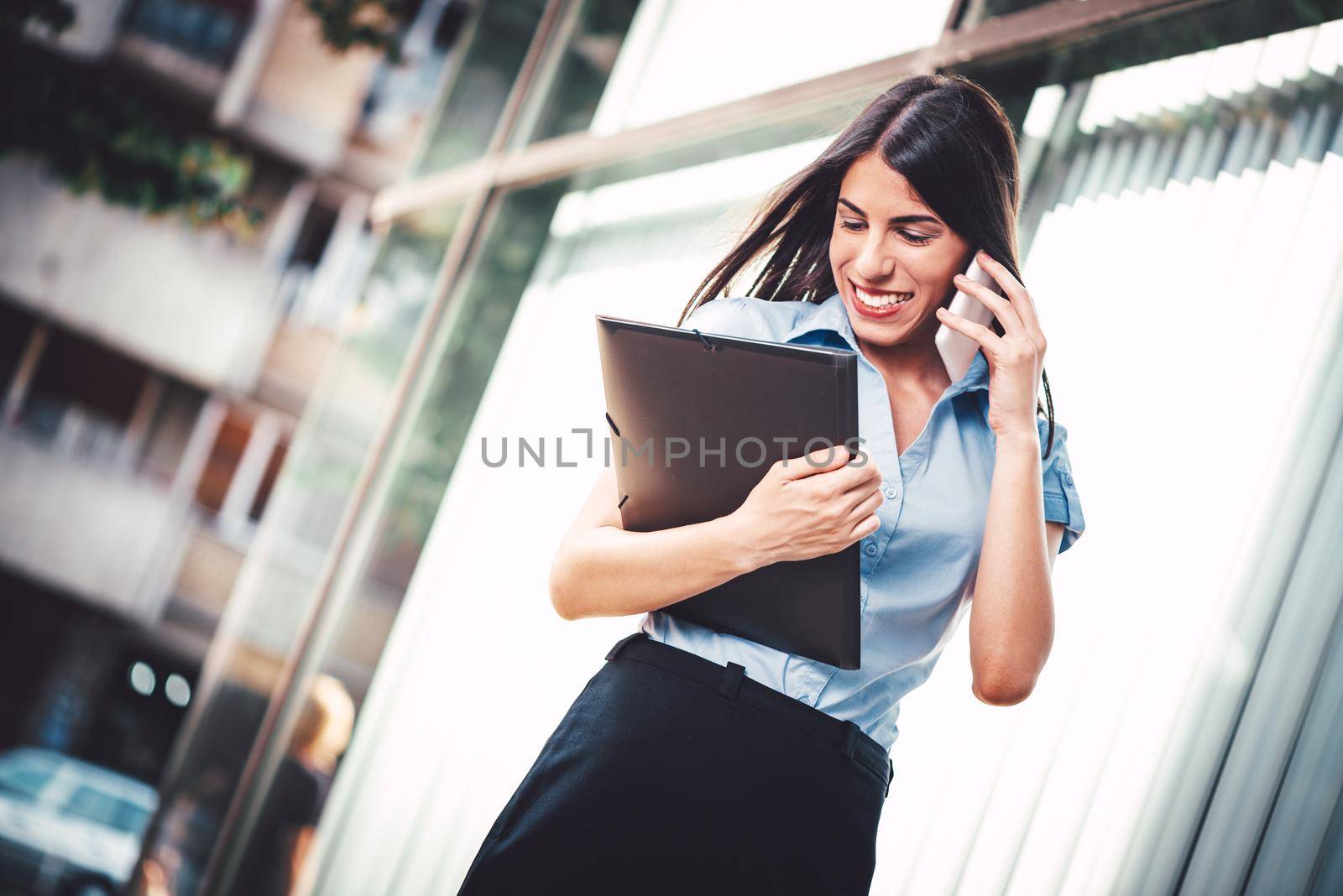 Emotional portrait of a happy and beautiful young businesswoman talking on a smartphone and reading information in a file document on the background of a business center.