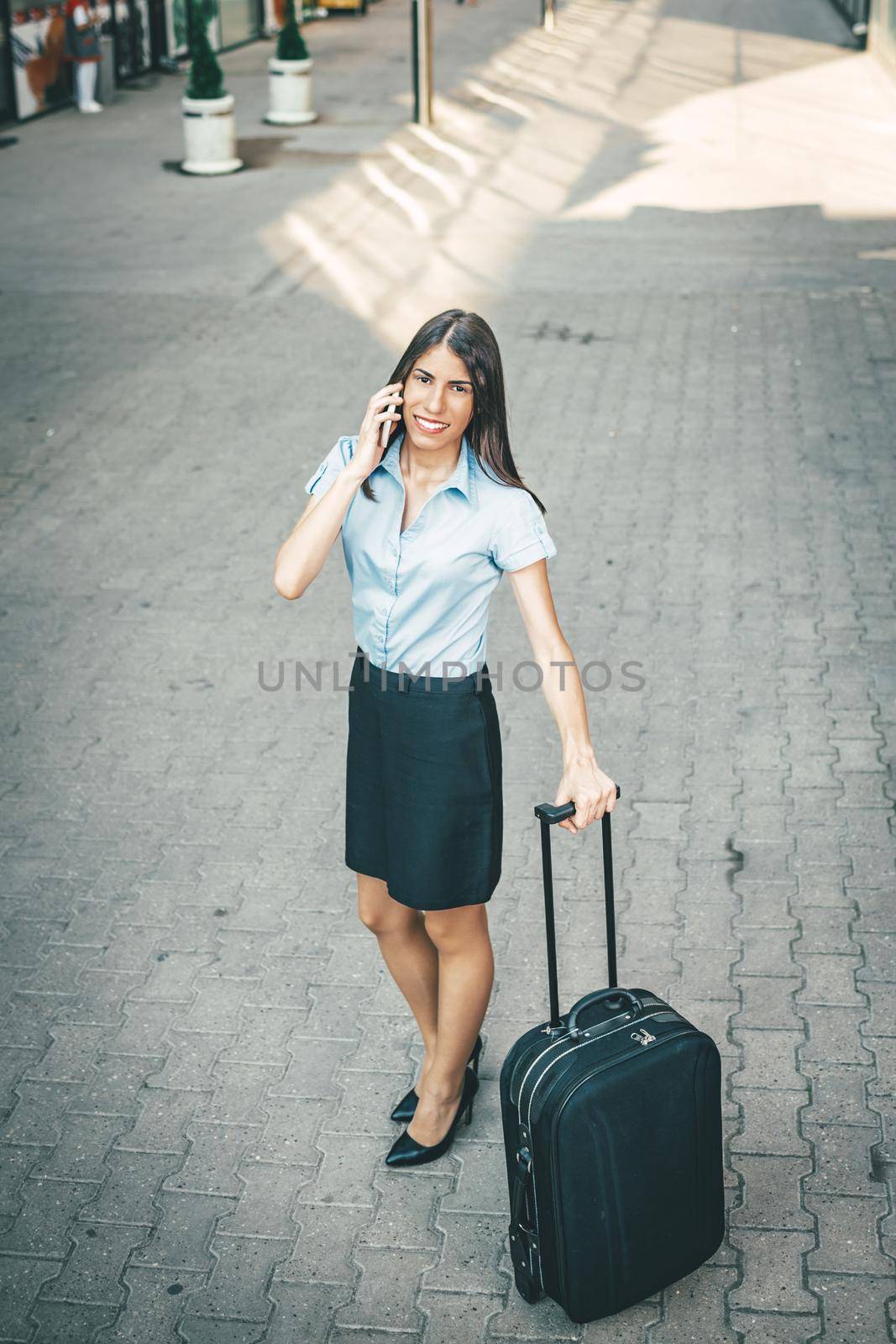 Young beautiful businesswoman standing outdoor with suitcase ready for a business trip and talking on mobile phone.