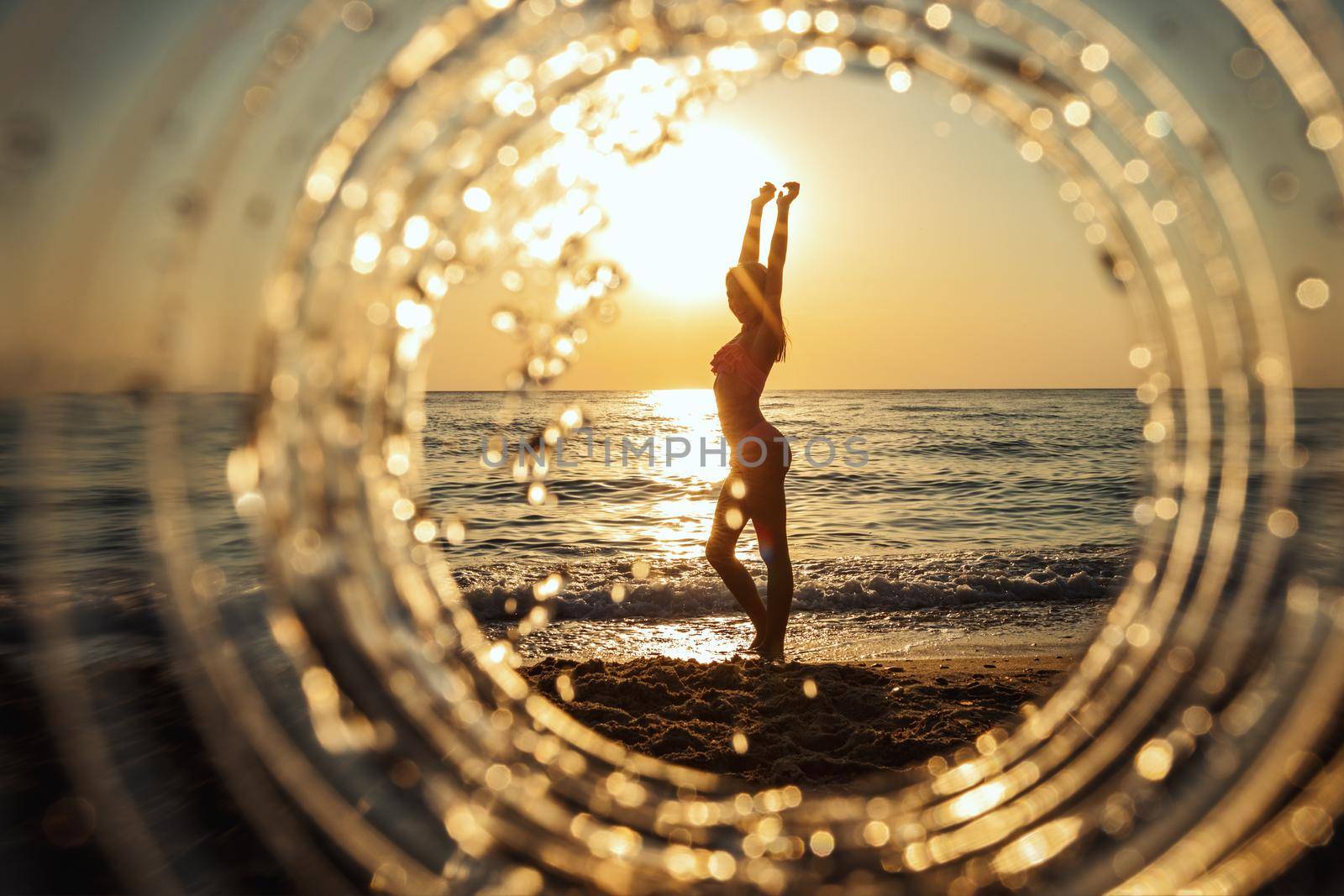 A beautiful creative composition of a sea landscape shot through a circle focus showing a teenage girl who posing on the beach in sunset.