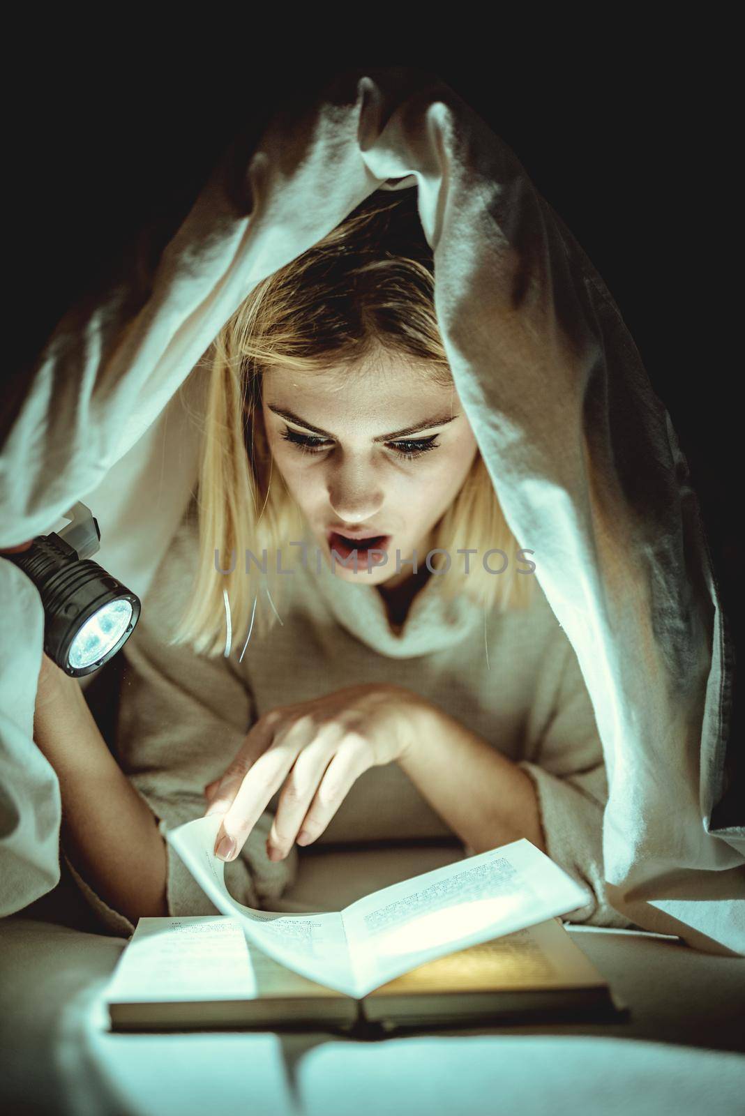 Beautiful young woman reading book under bed covering and holding a flashlight. 