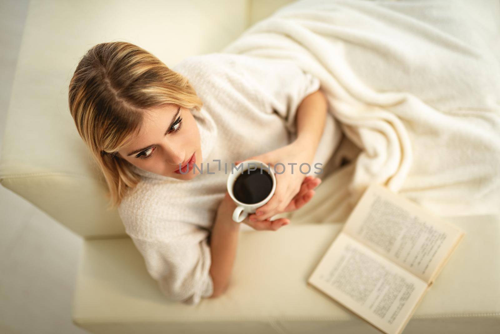 Beautiful young smiling woman is relaxing on her sofa in the living room drinking coffee and reading a book.