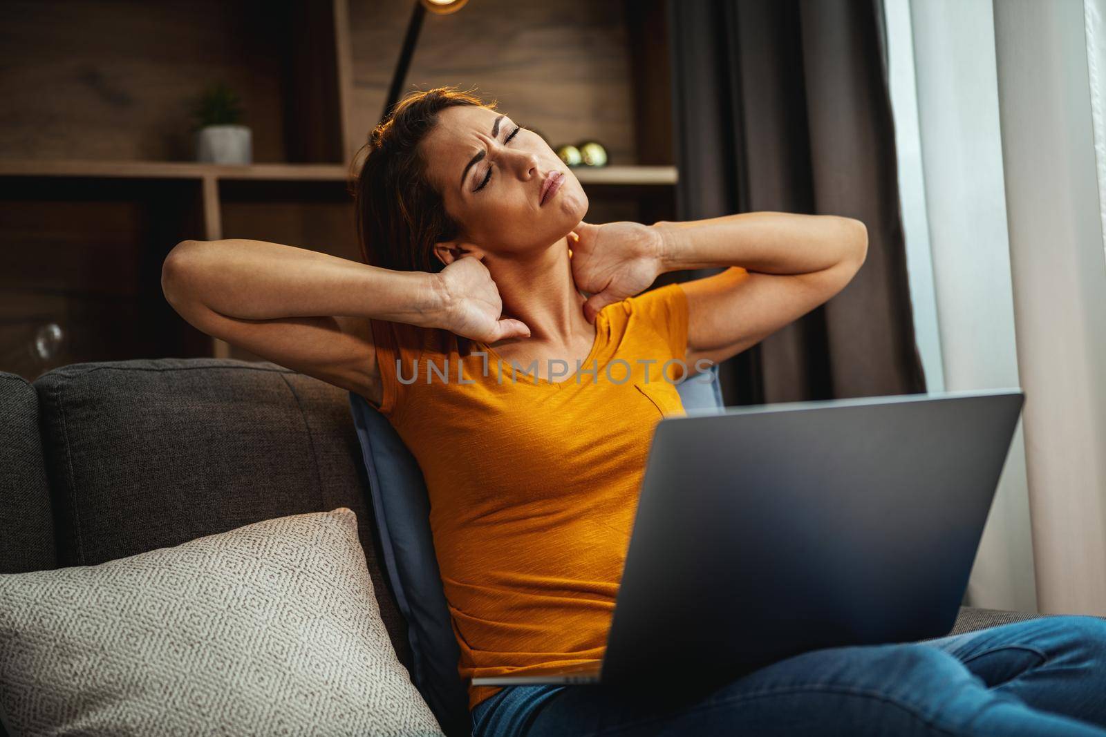 Shot of an tired young woman experiencing stress and holding her neck in pain while sitting on the sofa and using her laptop at home.