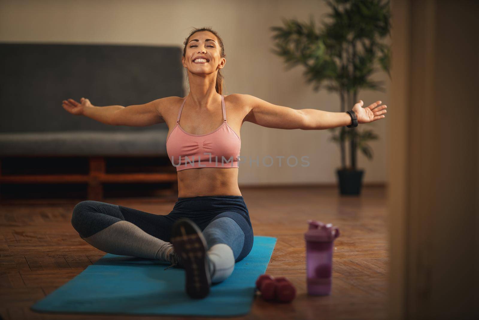 Young smiling woman is doing stretching exercises in the living room on floor mat at home in morning sunshine.