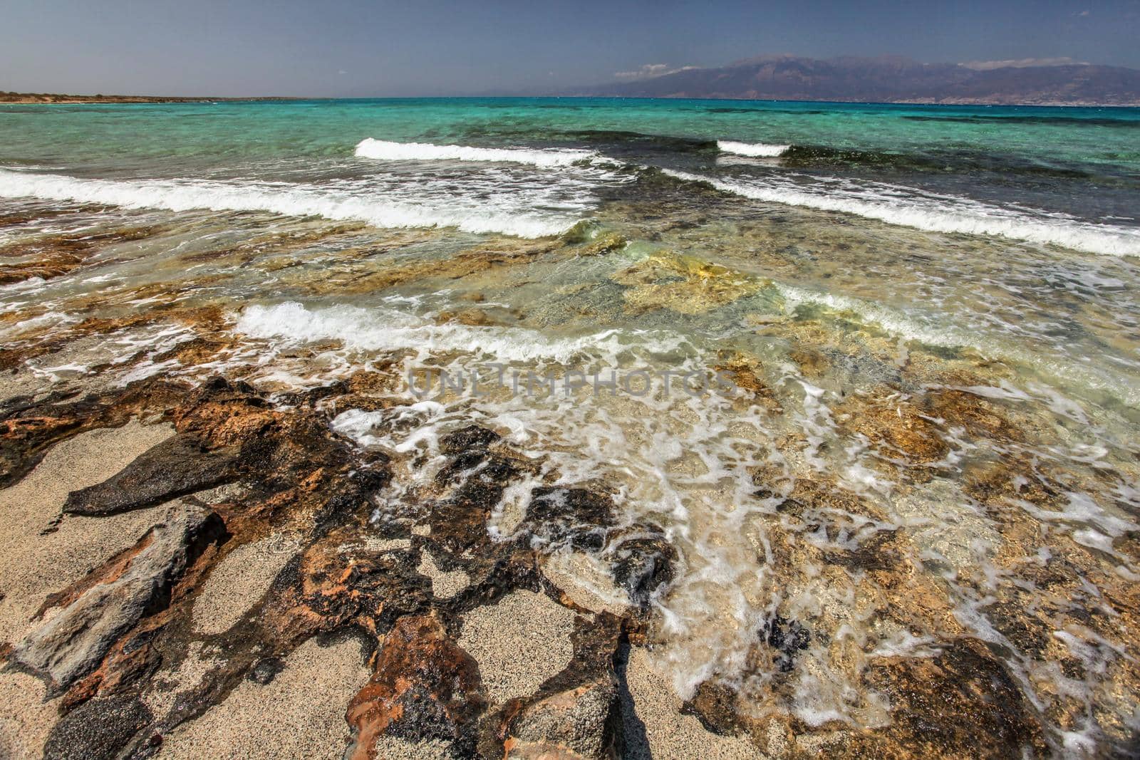 Sand on large rocks, crystal clear turquoise sea with white waves and land in background. Chrissi , Greece
