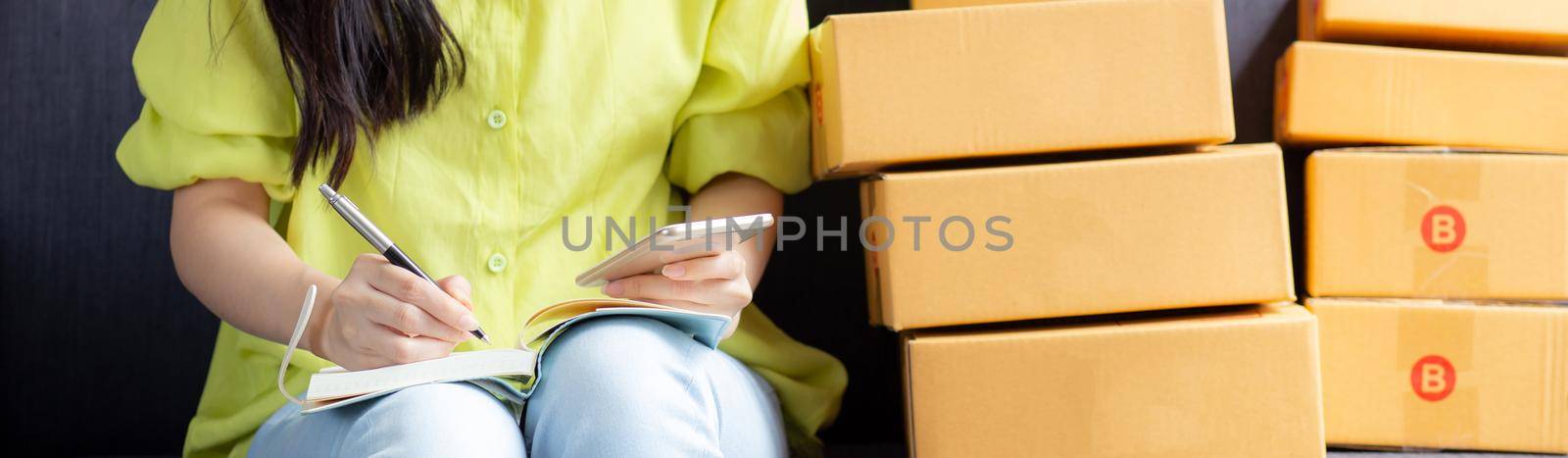 Young asian woman is entrepreneur looking phone and writing order of customer with online on notebook at home, online shopping, logistic and delivery, female is seller working on sofa, business SME.