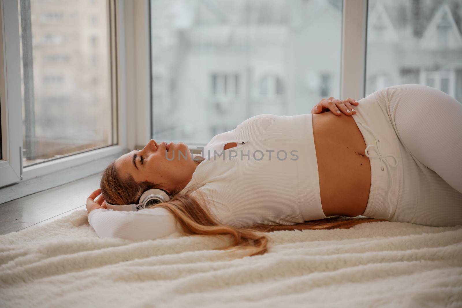 Side view portrait of relaxed woman listening to music with headphones lying on carpet at home. She is dressed in a white tracksuit. by Matiunina