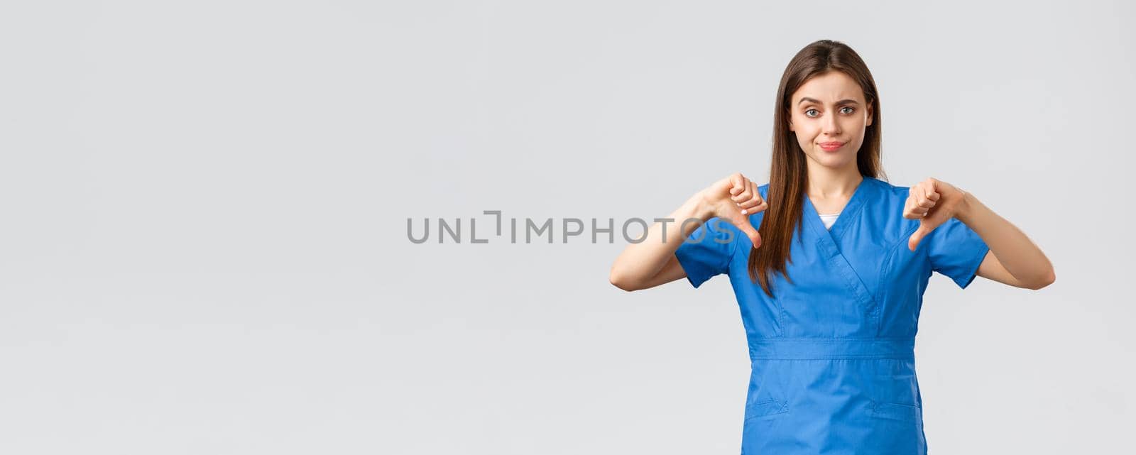 Healthcare workers, prevent virus, insurance and medicine concept. Skeptical and upset nurse or doctor in blue scrubs, smirk disappointed, showing thumbs-down dislike something.