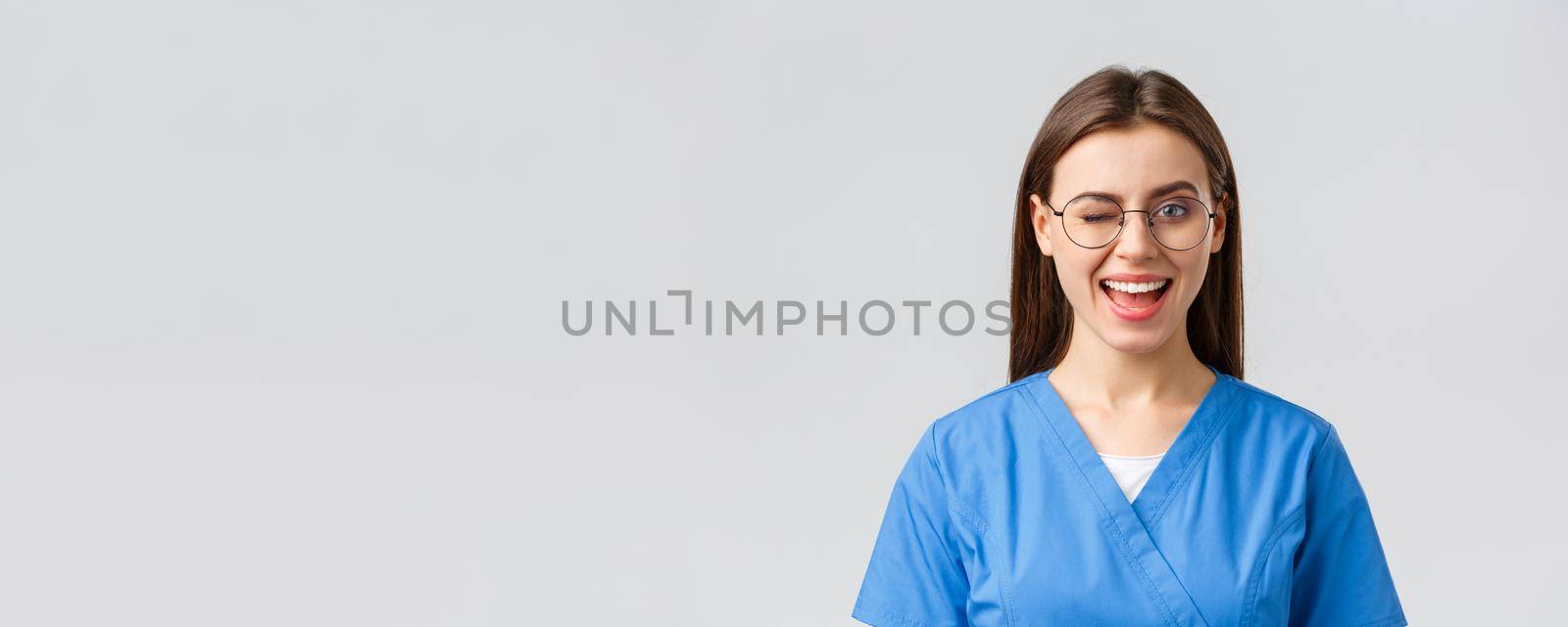 Healthcare workers, medicine, insurance and covid-19 pandemic concept. Cheeky pretty female nurse, intern or doctor in clinic, wear scrubs and glasses, wink at camera with enthusiastic smile.