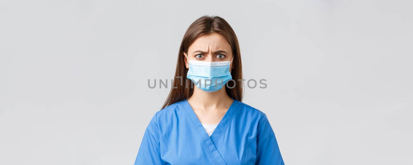 Covid-19, preventing virus, health, healthcare workers and quarantine concept. Angry or frustrated female nurse reacting to bad news. Doctor in blue scrubs and medical mask frowning disappointed.