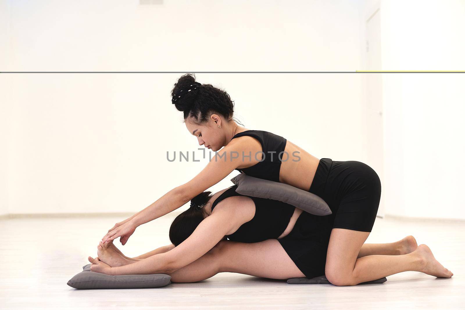 Young yoga female instructor assisting a beginner balancing flexibility pose in class at gym studio. Yoga and pilates classroom.