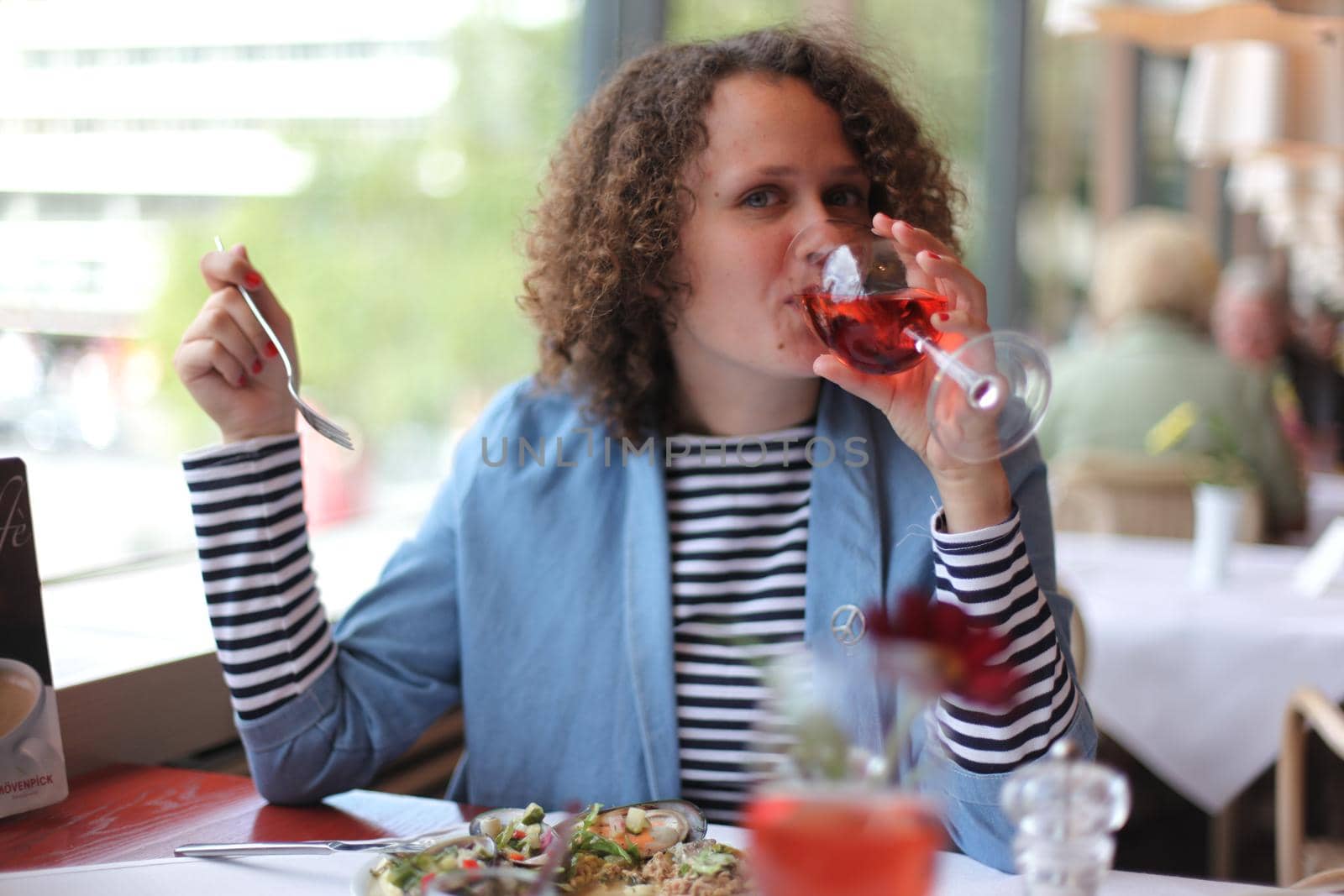 Happy stylish young female in casual outfit drinking wine and looking at camera while sitting at table during lunch in restaurant