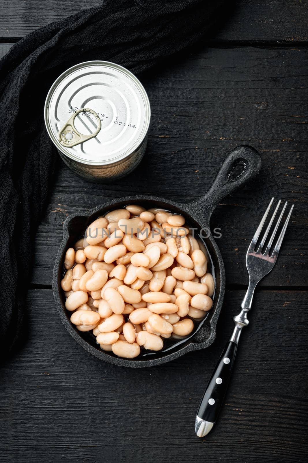 White kidney beans set, with metal can, in cast iron frying pan, on black wooden table background, top view flat lay