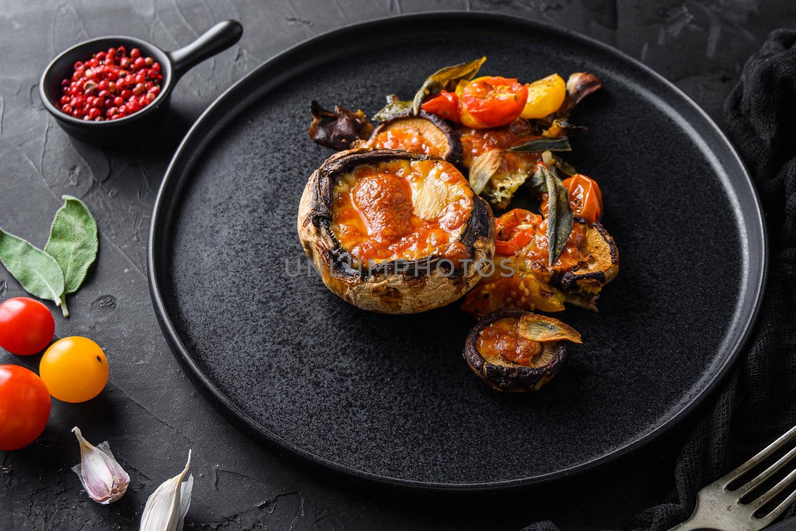 Served portobello mushrooms,baked and stuffed with cheddar cheese, cherry tomatoes and sage on black plate over black background side view