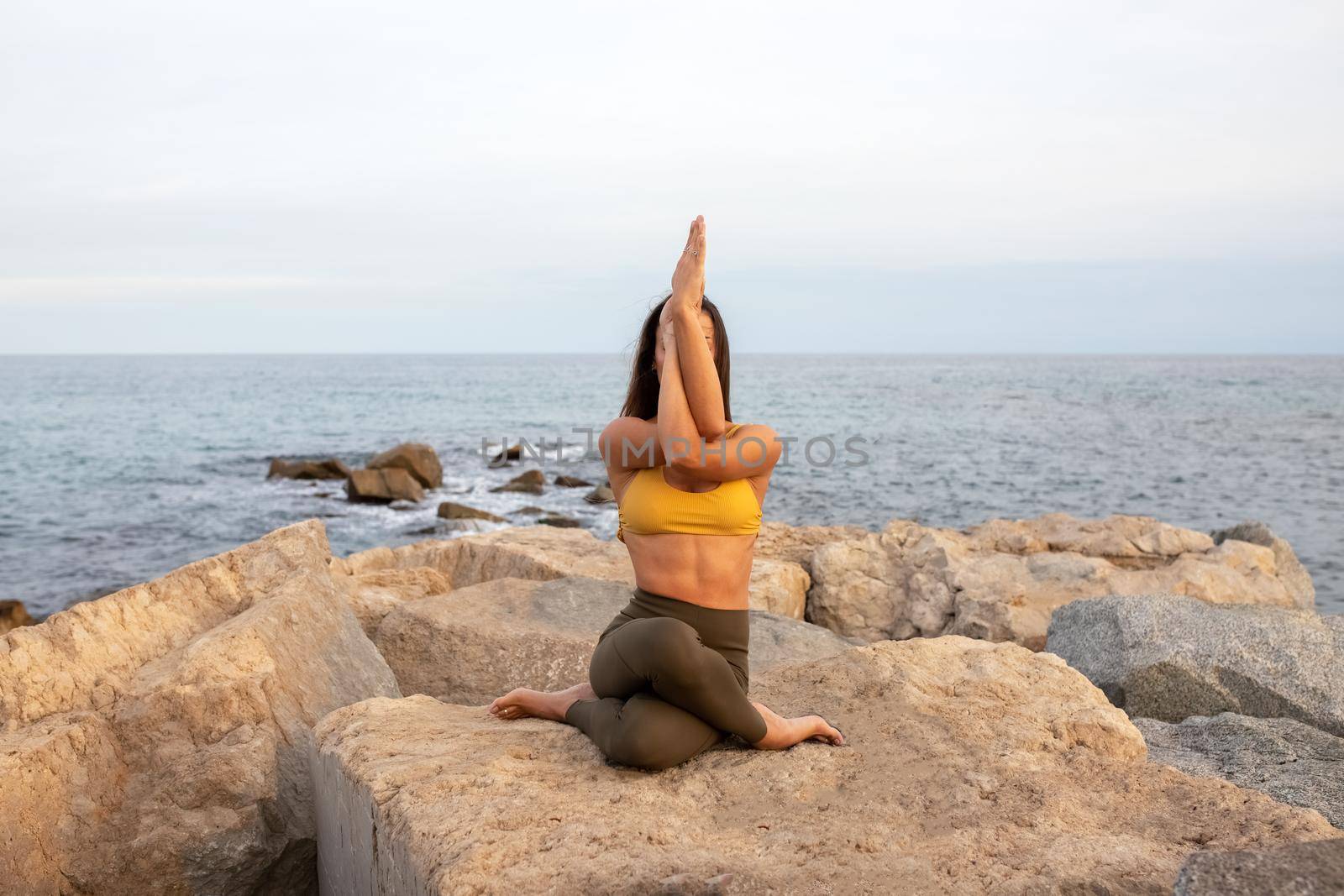 Unrecognizable woman doing gomukhasana with garudasana arms. Yoga practice in nature near the ocean. Healthy lifestyle and spirituality concept.
