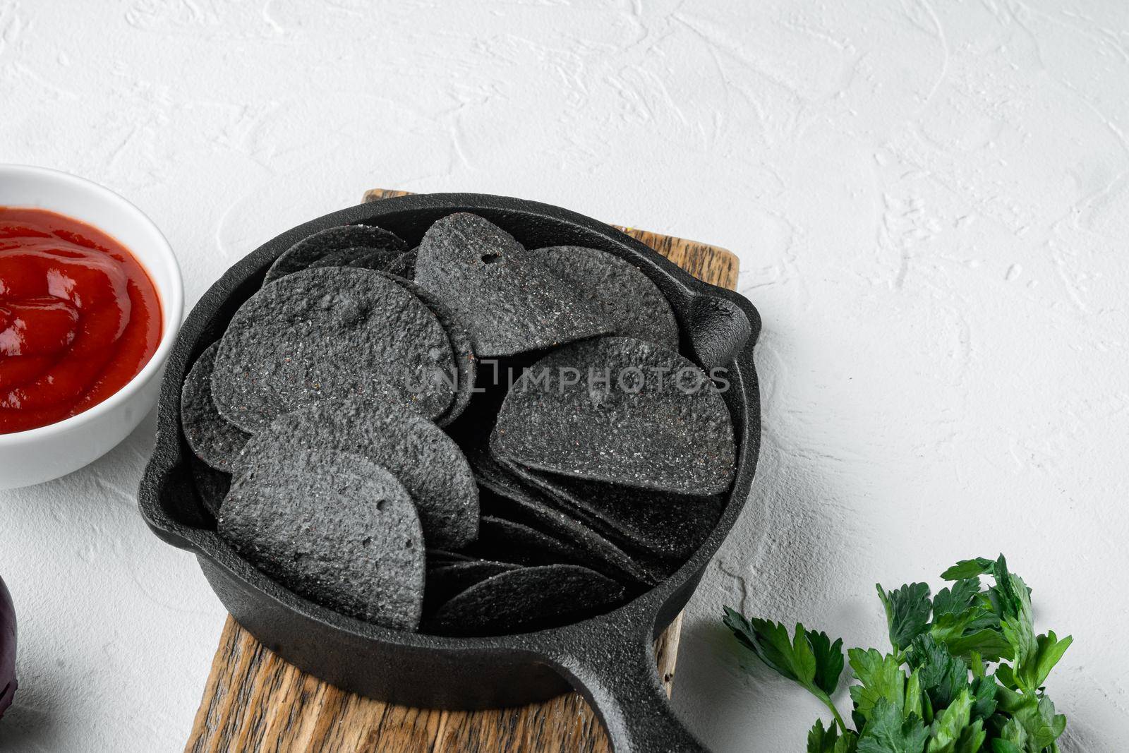 Black crispy potato chips set, with dipping sauces tomato dip sour cream, in cast iron frying pan, on white stone surface, with copy space for text