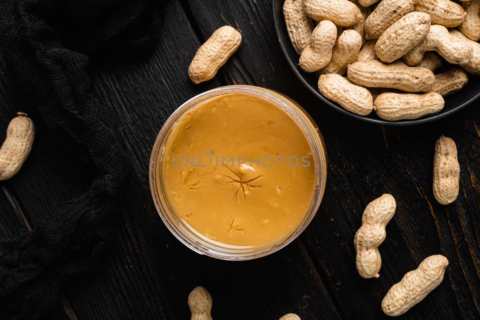 Peanut paste in an open jar and peanuts set, on black wooden table background, top view flat lay, with copy space for text