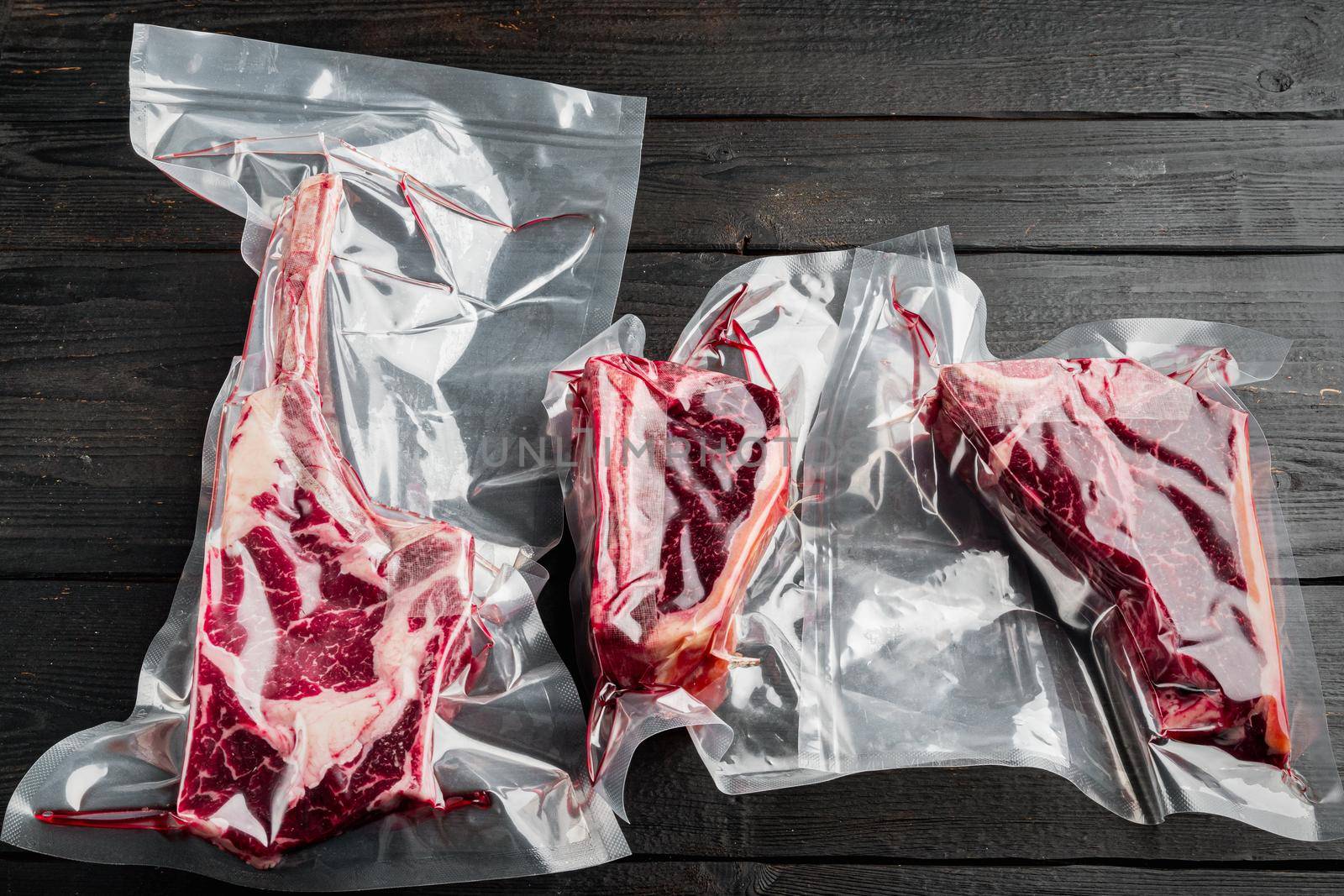 Dry aged steak in a vacuum. Meat products in plastic pack, tomahawk, t bone and club steak cuts, on black wooden table background by Ilianesolenyi