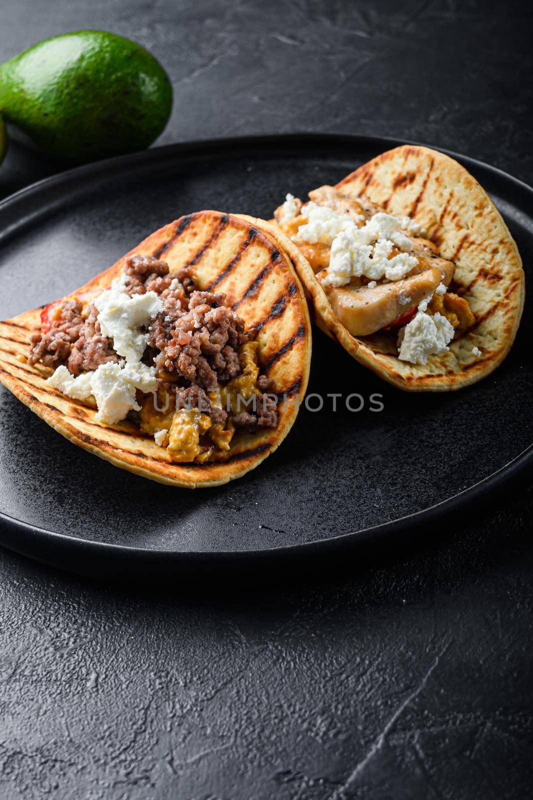 Mexican tacos with vegetables and beeaf and chicken meat on black round plate over textured black background, side view, selective focus vertical.