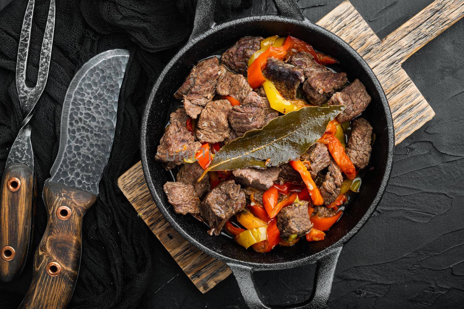 Beef meat stewed with potatoes, carrots and spices set, in cast iron frying pan, on black stone background, top view flat lay