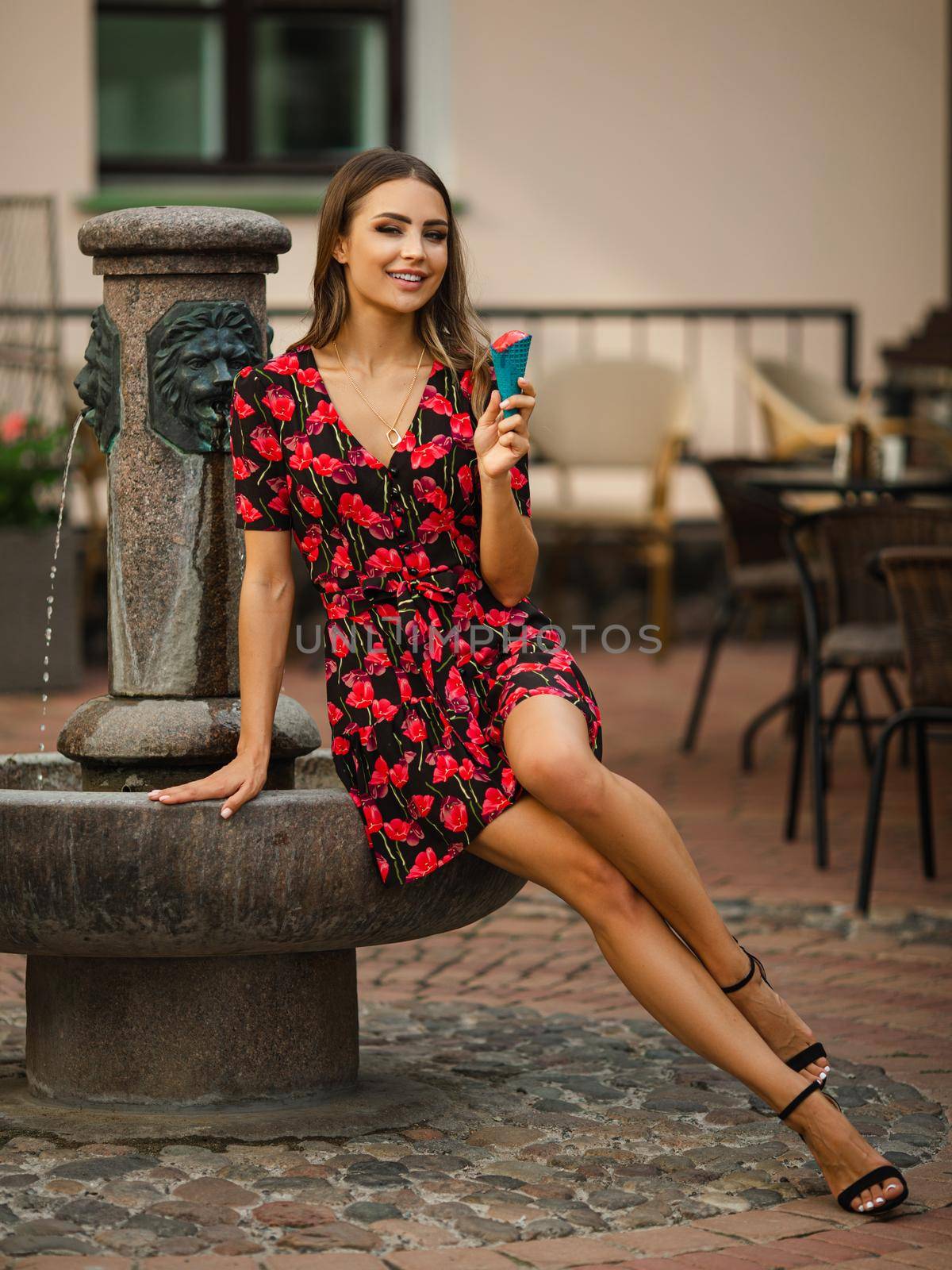 young cheerful woman with wavy hair wearing black dress with red flowers eating yummy ice-cream near the fountain by StudioLucky